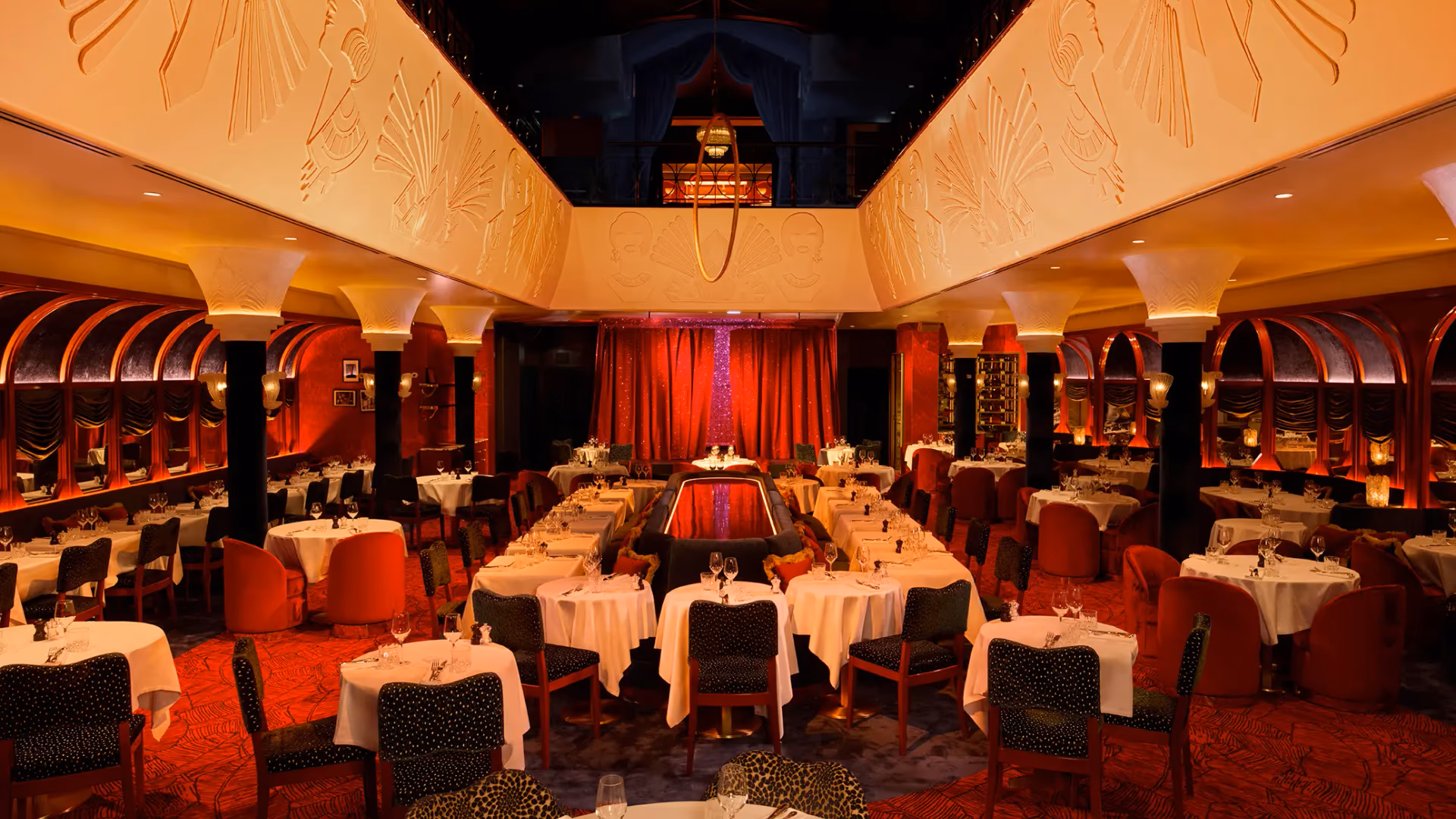 Salle de restaurant élégante avec des tables dressées, des chaises rouges et vertes, des colonnes noires et des murs décorés de rideaux rouges et noirs