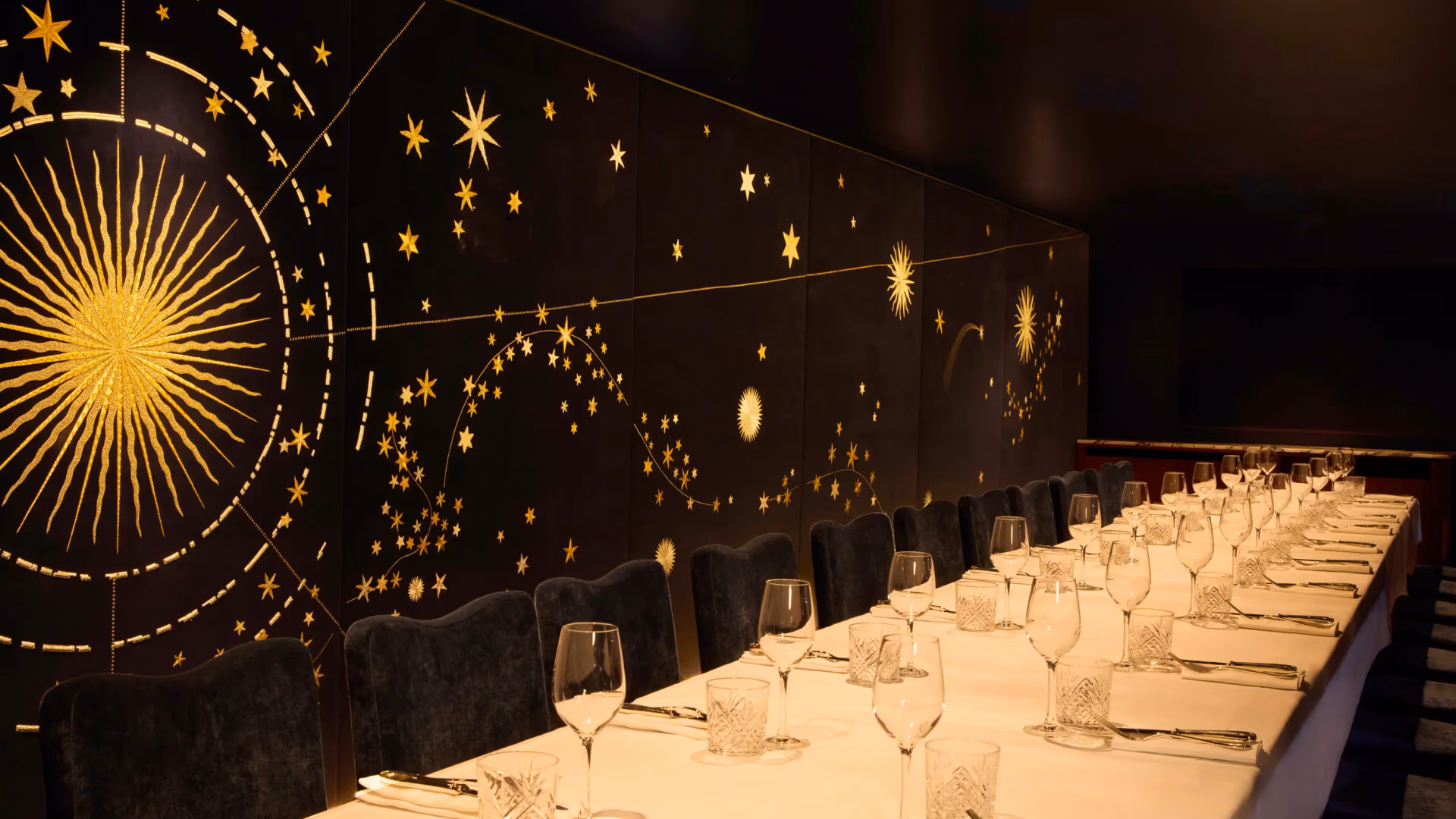 Une longue table dressée avec des verres et des couverts, entourée de chaises, dans une salle décorée de motifs étoilés lumineux sur fond noir